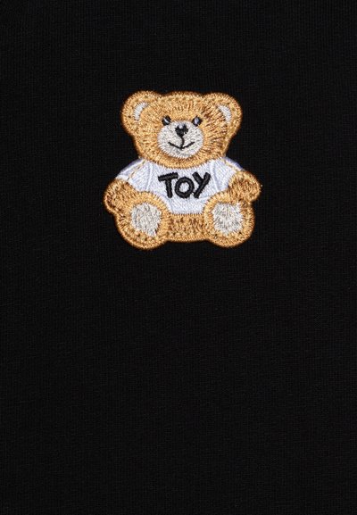 Ein schwarzer Stoff zeigt einen gestickten Teddy-Bären-Patch. Der Bär ist beige und trägt ein weißes Shirt mit der Aufschrift "TOY" in schwarzen Buchstaben.