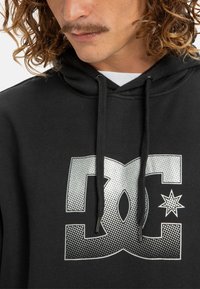 Čierna mikina s kapucňou s veľkým strieborným logom DC s bodkovaným vzorom a malým hviezdičkovým detailom. Vyrobená z jemného materiálu, doplnená o šnúrky.