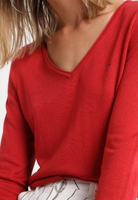 Roter Strickpullover mit V-Ausschnitt und langen Ärmeln. Mit einem kleinen Logoakzent auf der linken Seite. Kombiniert mit gestreiften weißen Hosen.