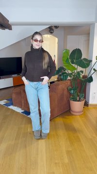 Rjava rebrasta pulover s turtleneck ovratnikom, svetlo modri kavbojki, sivi gležnjarji in rjave sončne očala v notranjem okolju z rastlinami.