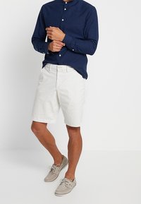 Man som bär en marinblå långärmad skjorta med knapprad, vita knälånga shorts och ljusbeiga båttofflor, stående mot en enfärgad vit bakgrund.