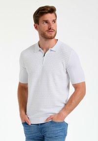 Polo de punto blanco con un patrón de diamantes en relieve, mangas cortas y cuello con cremallera. Llevado con unos pantalones vaqueros azules.