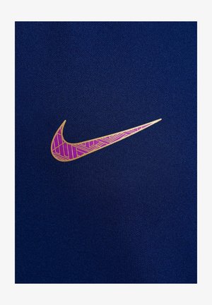 Logo Nike în aur, cu modele geometrice roz, pe un fundal de țesătură texturată de culoare albastru închis. Logo-ul are un design curbat, în formă de swoosh.