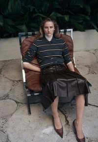 Falda plisada de cuero marrón, suéter a rayas azul marino y tacones puntiagudos marrones. Sentada en una silla acolchada marrón con estructura cromada.