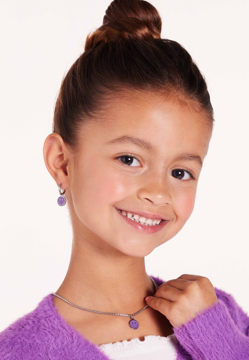 Pendientes de cara sonriente morados y colgante a juego en una cadena de plata. Suave y esponjoso suéter morado. El fondo brillante realza los colores de la joyería.
