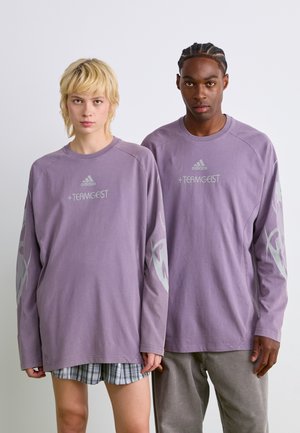 Chemises à manches longues, de couleur violet clair, avec un col rond. Présente le logo "adidas" et le texte "+ TEAMGEIST". Décorées de graphiques gris sur les manches.