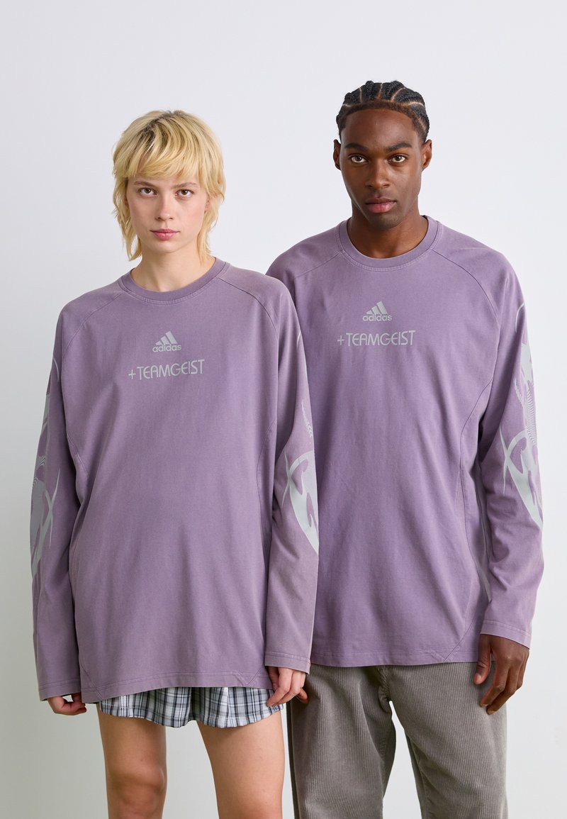 Chemises à manches longues, de couleur violet clair, avec un col rond. Présente le logo "adidas" et le texte "+ TEAMGEIST". Décorées de graphiques gris sur les manches.