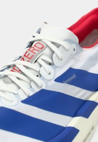 Weißer und blauer Sportschuh mit weißen Schnürsenkeln, rotem Innenfutter und rotem Schriftzug "N ZERO" auf der Zunge, seitliche und obere Details sichtbar.