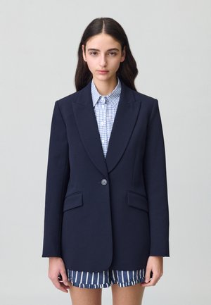 Blazer azul marino con solapas de muesca, cierre de un solo botón, dos bolsillos frontales, corte ajustado, combinado con una camisa a cuadros y pantalones cortos a rayas.