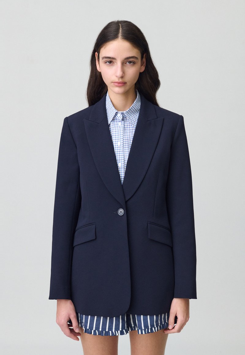 Blazer marine à revers crantés, fermeture à un bouton, deux poches avant, coupe ajustée, associé à une chemise à carreaux et un short rayé.