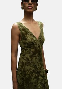 Vestido de terciopelo verde oliva con un escote en V profundo, silueta ajustada, patrón floral texturizado y sin mangas. Se aprecian accesorios dorados.