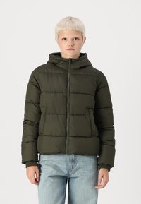 Maslinasto zelena puffer jakna s kapuljačom, zatvaranjem na munju i kviltanom teksturom. Ima bočne džepove i čvrste manžetne. Nosi se preko svjetloplavih traperica.