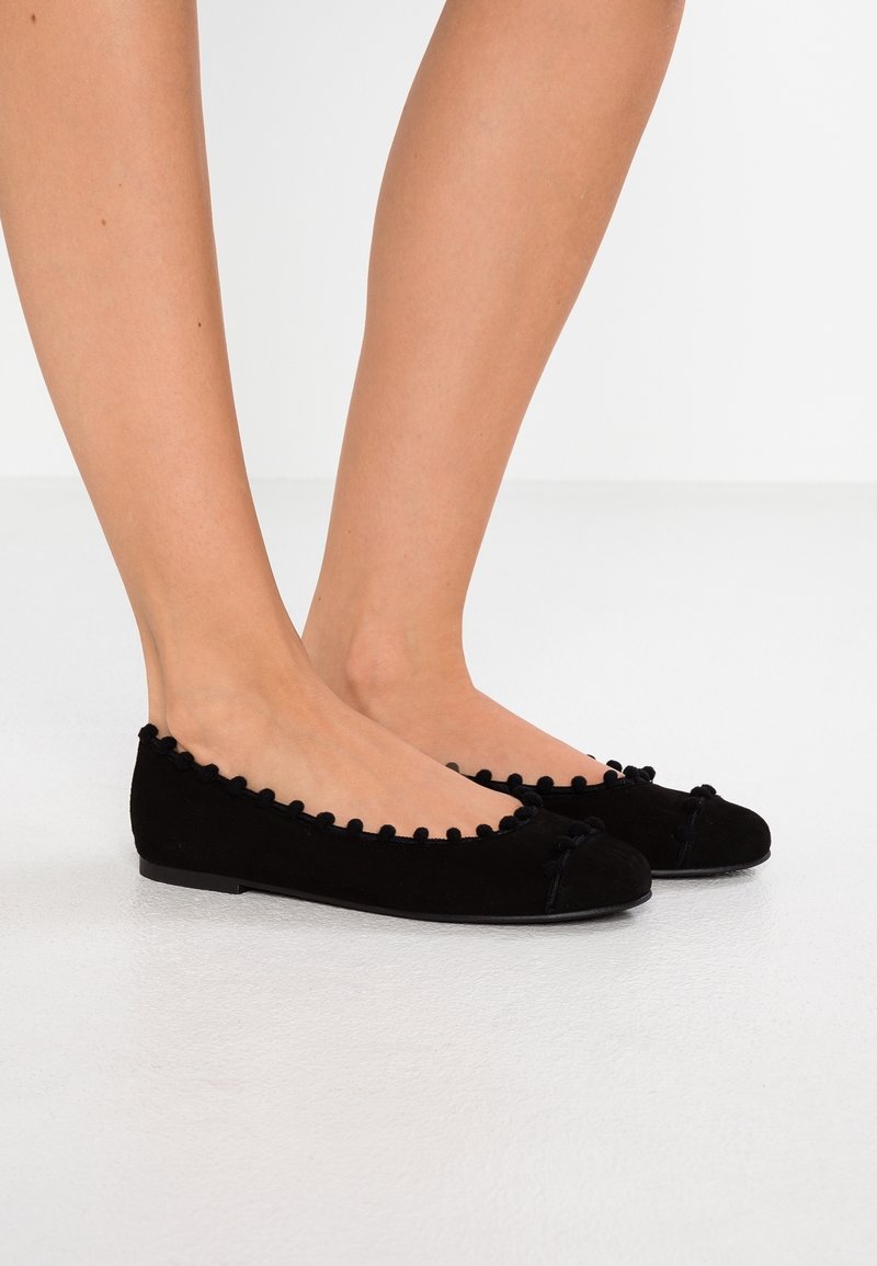 Ballerines en suede noir avec un bord à pompon décoratif, présentant un bout arrondi et un design minimaliste. Semelle plate, sans matériel visible.