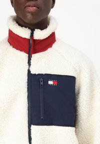 Giacca in pile crema con colletto rosso e blu, tasca con zip blu con logo, tessuto testurizzato e dettagli della zip visibili.