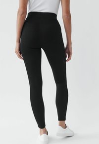 Zwarte high-waisted leggings met een gladde textuur, een strakke pasvorm en naadloze enkelmanchetten. Gecombineerd met witte schoeisel.