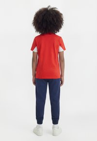 Παιδί με σγουρά μαλλιά που φοράει κόκκινο κοντομάνικο μπλουζάκι, σκούρο μπλε παντελόνι, λευκές κάλτσες και λευκά αθλητικά παπούτσια, στέκεται με την πλάτη στραμμένη.