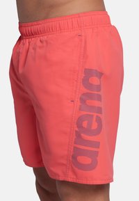 Costumi da bagno da uomo in un vivace corallo, realizzati in tessuto leggero. Presentano un cinturino elastico e un logo viola sul lato.