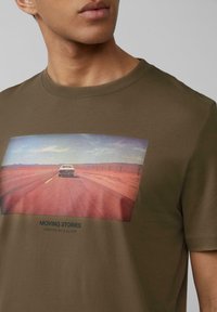 Olijfgroen katoenen t-shirt met een rechthoekige grafiek met een auto op een rode weg, de tekst "MOVING STORIES" erboven en "CRAFTED BY S.OLIVER" eronder.