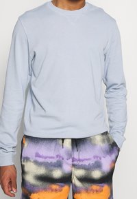 Světle modrý svetr s kulatým výstřihem, dlouhými rukávy a hladkou texturou, doplněný o vzorované vícebarevné šortky s fialovými a oranžovými akcenty.