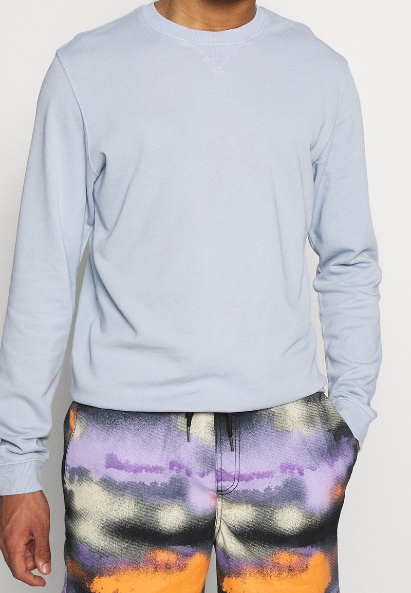 Světle modrý svetr s kulatým výstřihem, dlouhými rukávy a hladkou texturou, doplněný o vzorované vícebarevné šortky s fialovými a oranžovými akcenty.