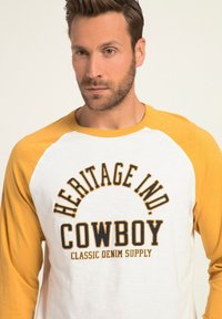 Witte t-shirt met gele raglanmouwen. Bevat de tekst "HERITAGE IND COWBOY CLASSIC DENIM SUPPLY" in zwarte en oranje letters op de voorkant.