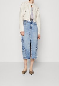 Chaqueta corta de cuero blanca con bolsillos con cremallera, combinada con una falda midi de mezclilla azul con una abertura frontal y bolsillos laterales tipo cargo. Zapatos planos marrones.