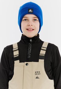 Zwarte met rits gesloten basislaag, beige sneeuwoverall met verstelbare schouderbanden en een blauwe gebreide muts met zwart logo-embleem.