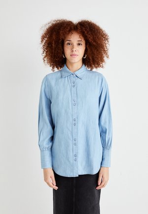 Camicia di denim azzurro chiaro con bottoni frontalmente, colletto classico e maniche lunghe con polsini arricciati. Texture liscia e design casual.
