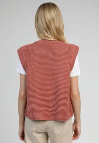 Haut sans manches en maille de couleur rose pâle, présentant un motif de point texturé, un col arrondi et un tissu doux. Porté sur une chemise blanche.