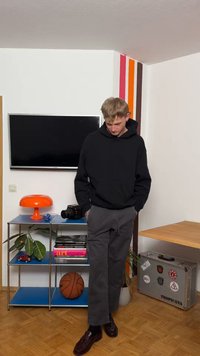 Νέος άνθρωπος με μαύρο φούτερ με κουκούλα και γκρι παντελόνι στέκεται σε ένα δωμάτιο με τηλεόραση, πορτοκαλί λάμπα, μπάλα του μπάσκετ, βιβλία και ένα διακοσμημένο μεταλλικό βαλιτσάκι.