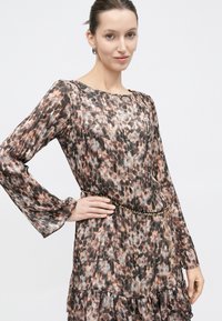 Νεαρή γυναίκα με μακρυμάνικο, πλισέ φόρεμα με animal print και λεπτή μαύρη και χρυσή αλυσίδα-ζώνη, χαμογελώντας με τα μαλλιά της πιασμένα προς τα πίσω.