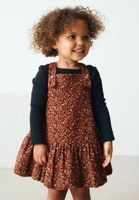 Kleid aus Corduroy mit Leopardenmuster und schwingendem Rock, verstellbaren Trägern und braunen Knöpfen, getragen über einem schwarzen Langarmshirt.