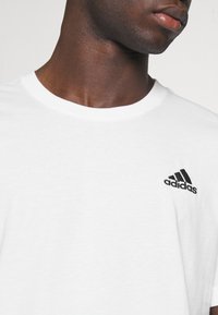 Bela bomba iz bombaža s krožnim ovratnikom in kratkimi rokavi, ki ima majhen črn vezen Adidas logotip na zgornjem levem prsnem delu.