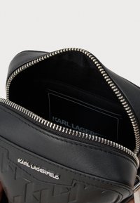 Melnas ādas rāvējslēdzēja soma ar teksturētu virsmu, sudraba tonis rāvējslēdzējs un iekšējā kabata ar karti, kas apzīmēta "KARL LAGERFELD".