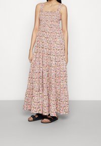 Robe maxi fleurie rose, à corsage smocké, encolure carrée et bretelles épaisses, avec une jupe à volants et des sandales noires.