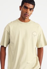 Light beige cotton t-shirt with short sleeves, a round neck, and a small cloud graphic on the chest. Soft texture, relaxed fit.