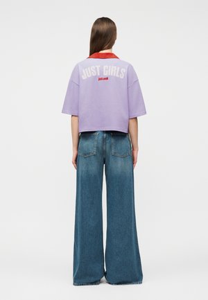 Vrouw met lang bruin haar draagt een lavendelgekleurde cropped top met een rode kraag waarop 'JUST GIRLS' staat, en wijde blauwe jeans, met de rug naar de camera toe.