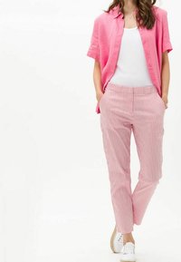Donna che indossa una camicia a maniche corte rosa con bottoni sopra un top bianco, pantaloni a righe rosa e bianchi e sneaker bianche, in piedi con le mani nelle tasche.