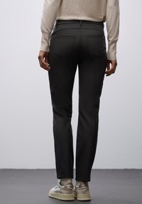 Osoba w czarnych spodniach, beżowym swetrze i białych trampkach, stojąca na szarym podłożu na tle jednolitej jasnoszarej ściany.