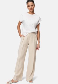 Chemise blanche à manches courtes avec des embellissements subtils, associée à un pantalon beige ample et des sandales plates avec une bride décorative.