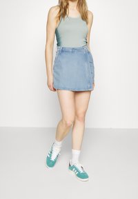 Mujer vestida con una blusa sin mangas de color azul claro, falda envolvente de mezclilla, calcetines blancos y zapatillas de deporte en color azul verdoso, caminando frente a un fondo blanco plano.