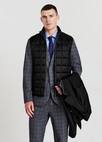 Homme en costume à carreaux gris et cravate bleue portant un gilet matelassé noir et tenant une veste noire contre un fond blanc uni.