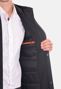 Giacca esterna nera con zip frontale, materiale leggero e tasca interna con accenti arancioni. L'interno presenta un rivestimento trapuntato.