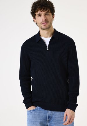 Sweater de punto navy con cuello con cremallera, diseño texturizado de canalé, mangas largas y dobladillo ajustado. Lleva puesto una camiseta blanca.
