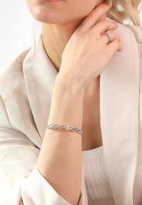 Silberner geflochtener Armband mit verflochtenen Strängen, der eine glatte Textur und ein poliertes Finish aufweist. Am nackten Handgelenk neben einem weichen Stoffärmel getragen.