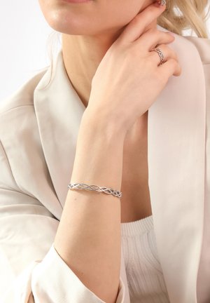 Silver braided bracelet with intertwined strands, featuring a smooth texture and polished finish. Worn on a bare wrist beside a soft fabric sleeve.