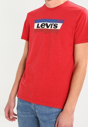 Czerwony bawełniany t-shirt z okrągłym dekoltem, z białym, niebieskim i czerwonym logo Levi's z przodu. Noszony z niebieskimi dżinsami.