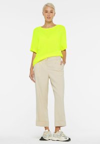 Neon-gelbes, kurzärmeliges Netzoberteil, das mit beigefarbenen, maßgeschneiderten Hosen kombiniert wird. Die Hosen haben umgeschlagene Säume, die durch helle Sneakers ergänzt werden.