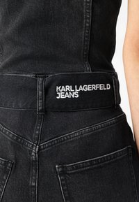 Juodos džinsinės kelnės turi ryškų juosmens etikėtę su „KARL LAGERFELD JEANS“ balta siuvinėjimu. Dizainas apima matomą siuvimą ir kišenes.