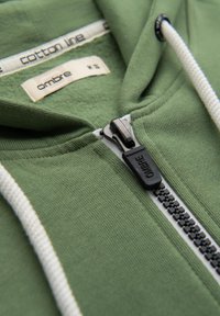 Olivenfarvet hættejakke lavet af bomuld, med en lynlås foran, hvide snore og en tekstureret inderside. Etikettedetaljer er synlige indeni.